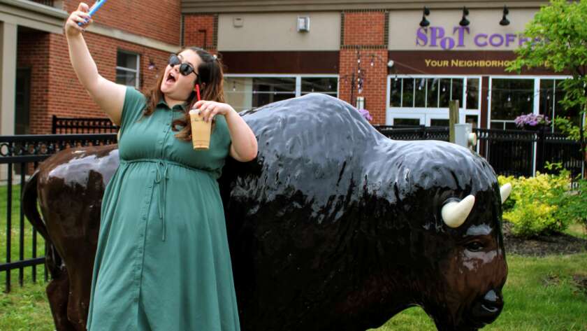 A photo of Kenmore Media Owner Rebecca Nagel posing to take a selfie with the buffalo statue in the village of Kenmore New York. She is wearing a teal dress and is holding an iced vanilla latte from Red Otter Coffee