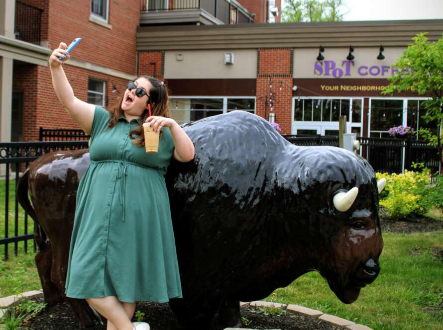 A photo of Kenmore Media Owner Rebecca Nagel posing to take a selfie with the buffalo statue in the village of Kenmore New York. She is wearing a teal dress and is holding an iced vanilla latte from Red Otter Coffee